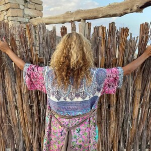May include: A woman wearing a long, multi-colored patterned robe with a brown sash tied at the waist. The robe has a floral print and a paisley design. The woman is standing in front of a woven bamboo fence.