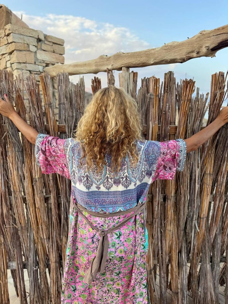 May include: A woman wearing a long, multi-colored patterned robe with a brown sash tied at the waist. The robe has a floral print and a paisley design. The woman is standing in front of a woven bamboo fence.