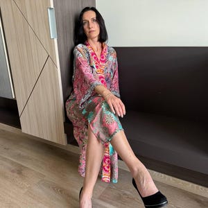 May include: A woman wearing a long, pink, and green patterned robe with a pink and orange trim. She is sitting on a dark brown bench with her legs crossed and wearing black heels.