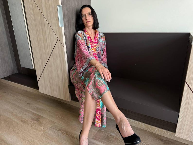 May include: A woman wearing a long, pink, and green patterned robe with a pink and orange trim. She is sitting on a dark brown bench with her legs crossed and wearing black heels.
