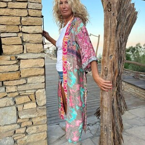 May include: A woman wearing a long, pink and blue patterned kimono with a white t-shirt underneath. She is standing in front of a stone wall and holding a piece of driftwood.