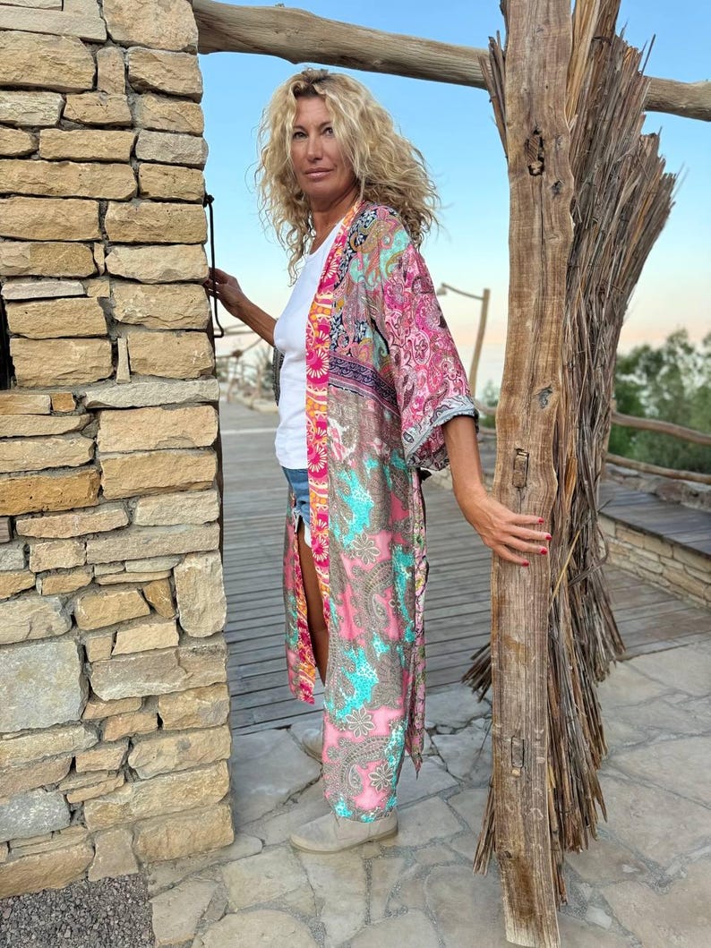 May include: A woman wearing a long, pink and blue patterned kimono with a white t-shirt underneath. She is standing in front of a stone wall and holding a piece of driftwood.
