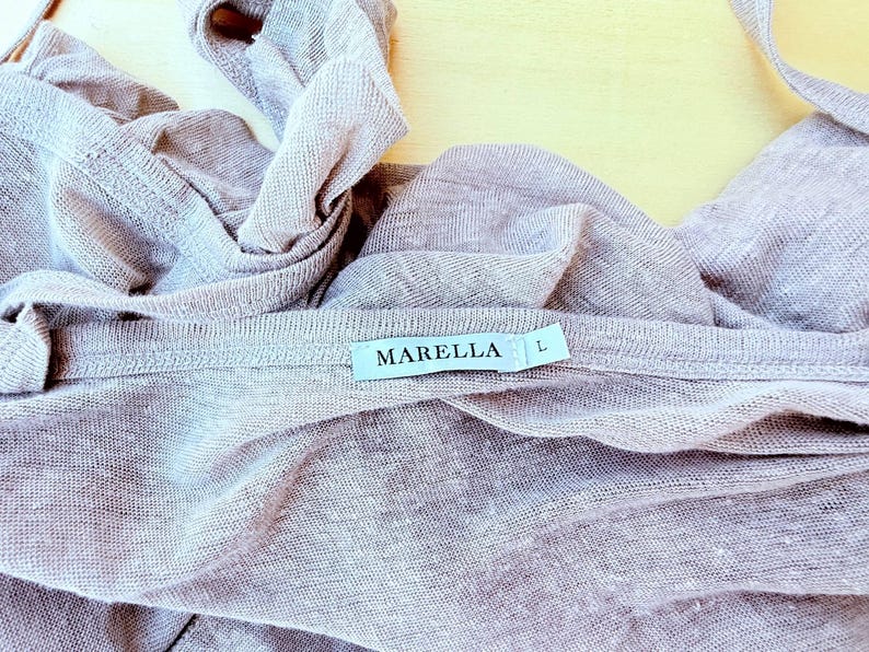 May include: A light mauve, semi-sheer knit garment featuring a white fabric label stitched at the neckline. The label displays the text MARELLA in black capital letters, followed by the letter L to indicate size. The fabric has a visible texture.