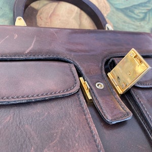 May include: A brown leather briefcase with a gold metal clasp. The briefcase has two pockets on the front and a handle on the top.