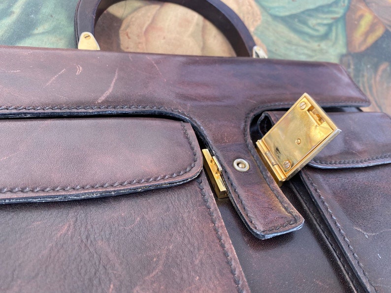 May include: A brown leather briefcase with a gold metal clasp. The briefcase has two pockets on the front and a handle on the top.