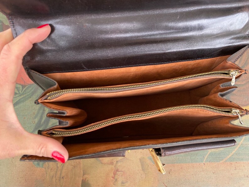 May include: A brown leather briefcase with two zippered compartments and a smaller pocket on the front. The interior is lined with a brown fabric.