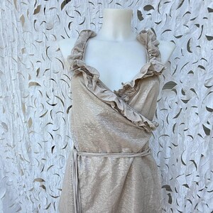 May include: A sleeveless wrap top in a shimmering metallic champagne gold fabric. The garment features a deep V-neckline framed by wide, ruffled fabric trim along the edges and a matching tie belt at the waist to secure the wrap-style closure.