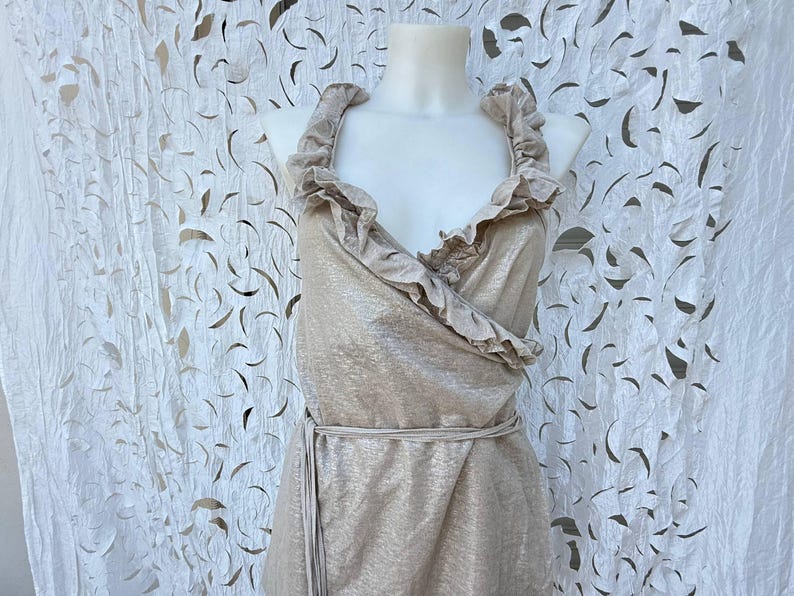 May include: A sleeveless wrap top in a shimmering metallic champagne gold fabric. The garment features a deep V-neckline framed by wide, ruffled fabric trim along the edges and a matching tie belt at the waist to secure the wrap-style closure.