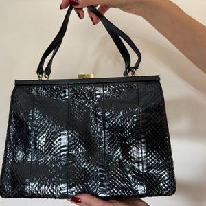 Può includere: Una borsa nera con motivo a pelle di serpente. La borsa ha una forma rettangolare, una chiusura a scatto e due manici neri. I dettagli metallici sono color oro.
