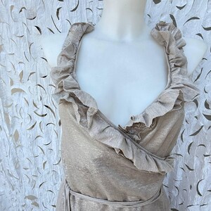 May include: A sleeveless wrap dress in a metallic champagne gold finish featuring a deep V-neckline. The bodice is accented with wide, ruffled fabric trim that follows the neckline and continues down the front, secured by a matching waist tie.