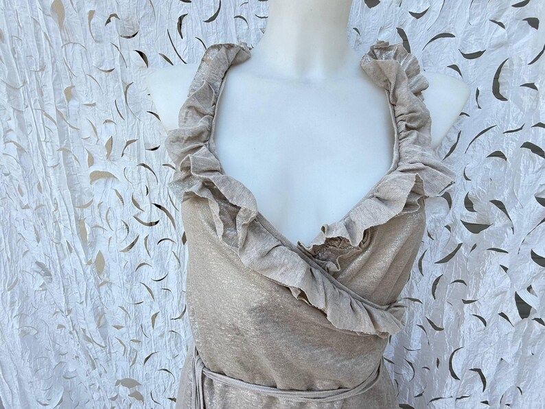 May include: A sleeveless wrap dress in a metallic champagne gold finish featuring a deep V-neckline. The bodice is accented with wide, ruffled fabric trim that follows the neckline and continues down the front, secured by a matching waist tie.