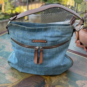 May include: A blue denim tote bag with brown leather straps and a zipper closure. The bag has a brown leather tag with the brand name on it.