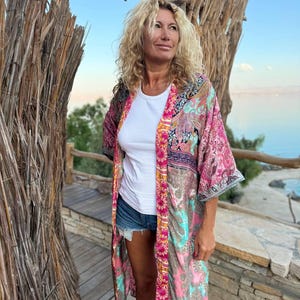 May include: A woman wearing a long, colorful patterned kimono with a white t-shirt and denim shorts. The kimono has a pink, turquoise, and gold color scheme with a paisley design.