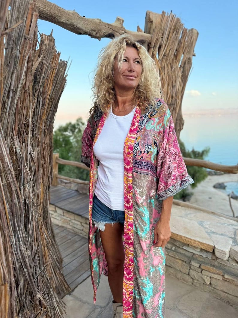 May include: A woman wearing a long, colorful patterned kimono with a white t-shirt and denim shorts. The kimono has a pink, turquoise, and gold color scheme with a paisley design.