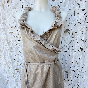 May include: A sleeveless wrap top in a metallic champagne gold finish, featuring a deep V-neckline with ruffled trim along the edges. The garment includes a thin, matching fabric tie at the waist and a straight hemline, displayed on a mannequin.