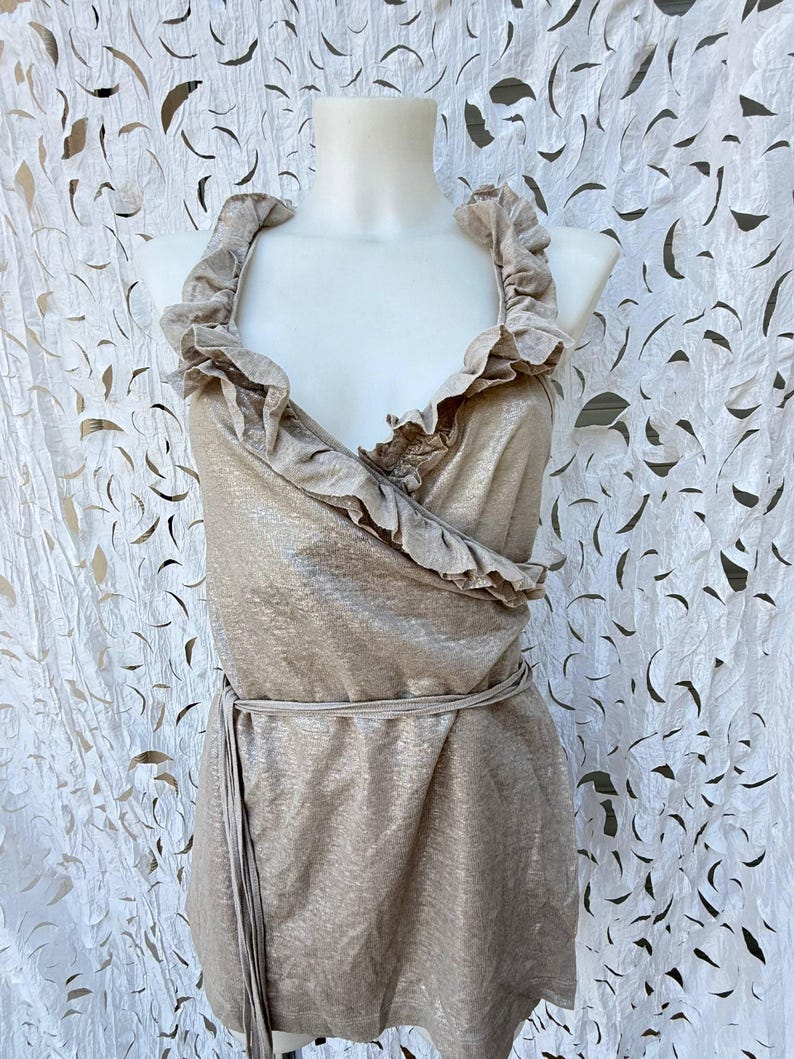 May include: A sleeveless wrap top in a metallic champagne gold finish, featuring a deep V-neckline with ruffled trim along the edges. The garment includes a thin, matching fabric tie at the waist and a straight hemline, displayed on a mannequin.