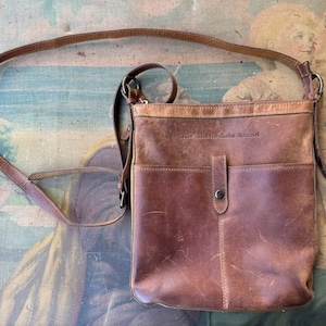 Puede incluir: Un bolso de cuero marrón cruzado con cierre de solapa y un bolsillo frontal. El bolso tiene una correa y el texto "The Sherpa Brand" está estampado en la parte delantera.