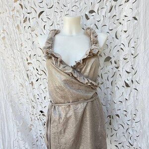 May include: A sleeveless metallic gold wrap top featuring a deep V-neckline, ruffled trim along the collar and front opening, and a thin matching fabric tie at the waist. The garment has a shimmering finish and a structured, fitted silhouette.