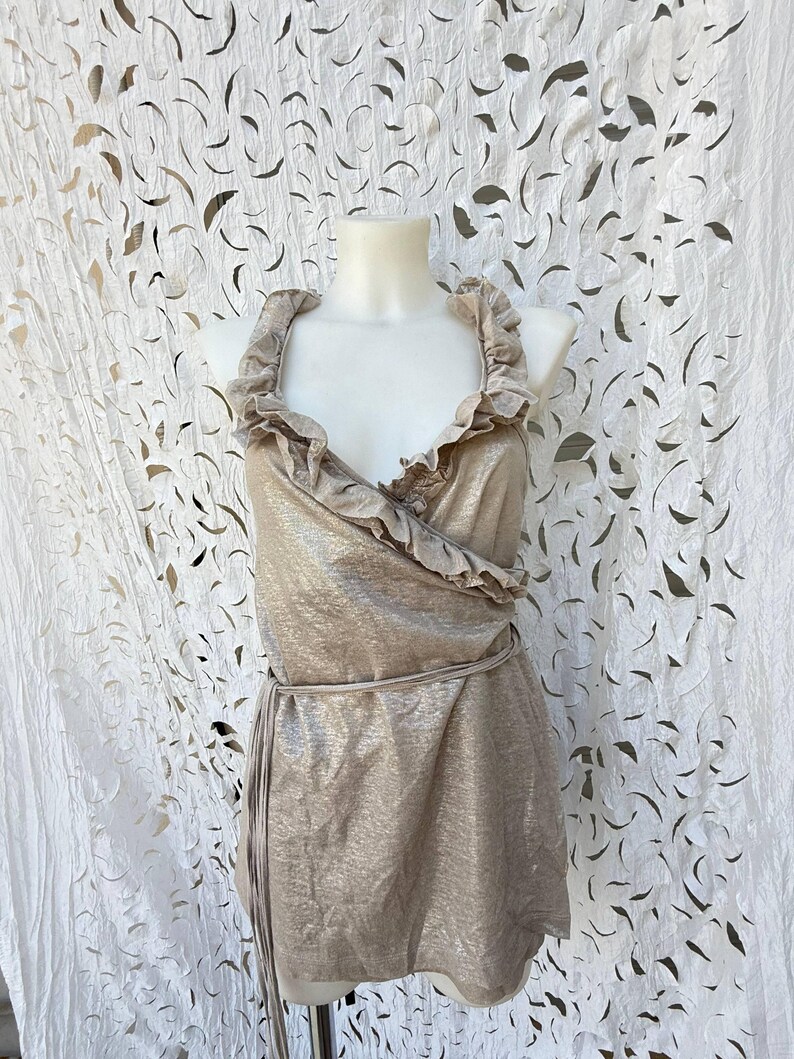 May include: A sleeveless metallic gold wrap top featuring a deep V-neckline, ruffled trim along the collar and front opening, and a thin matching fabric tie at the waist. The garment has a shimmering finish and a structured, fitted silhouette.