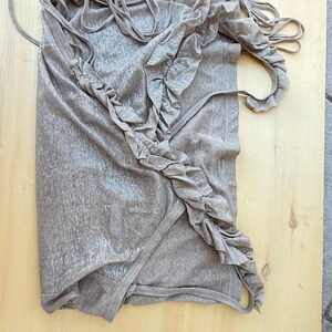 May include: A sleeveless top in a metallic taupe finish featuring a diagonal ruffled trim running from the shoulder to the hem. The garment includes multiple thin spaghetti straps at the top and a wrap-style front construction with a finished edge.