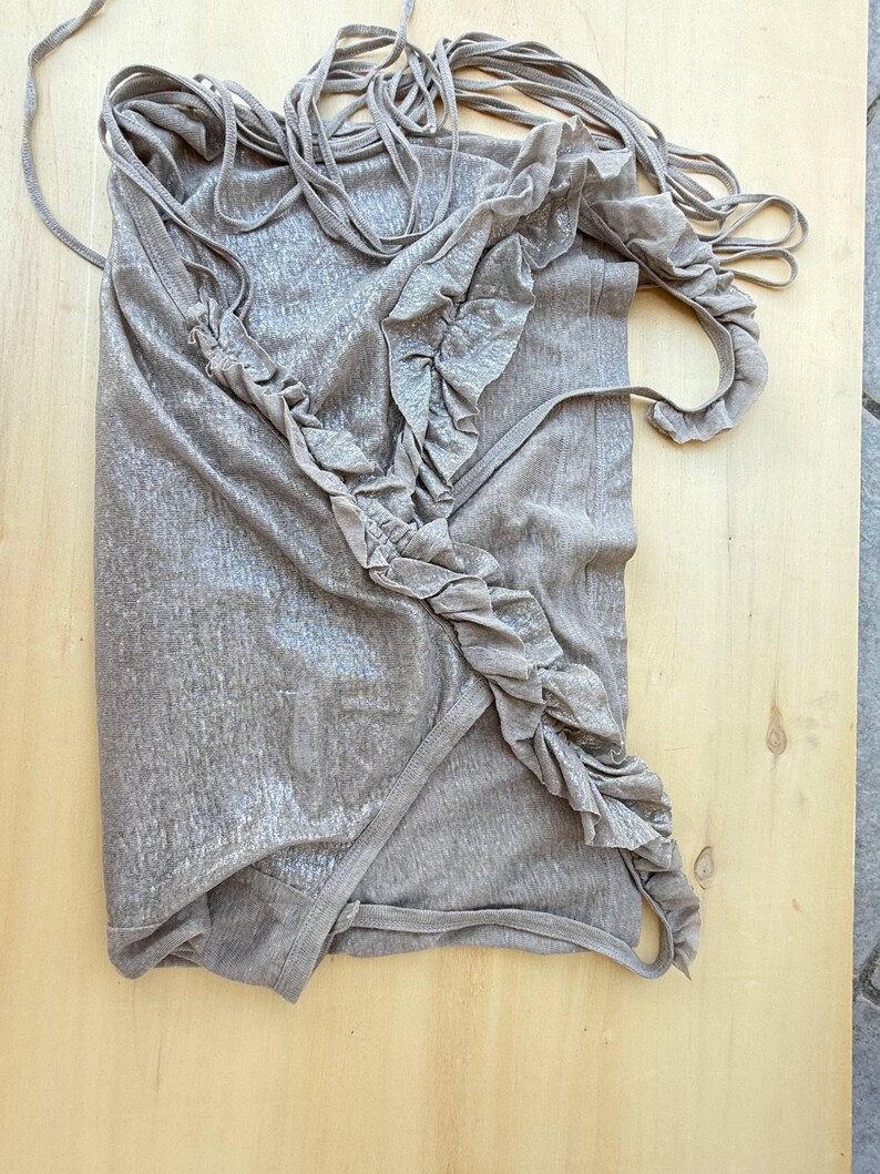May include: A sleeveless top in a metallic taupe finish featuring a diagonal ruffled trim running from the shoulder to the hem. The garment includes multiple thin spaghetti straps at the top and a wrap-style front construction with a finished edge.
