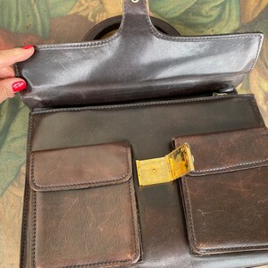 May include: A brown leather briefcase with a gold clasp and two front pockets. The briefcase has a top handle and a zipper closure.