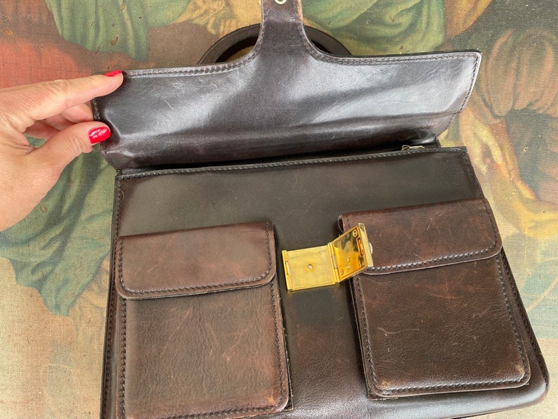 May include: A brown leather briefcase with a gold clasp and two front pockets. The briefcase has a top handle and a zipper closure.