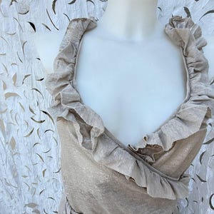 May include: A sleeveless beige top featuring a deep V-neckline and wide, ruffled trim that extends from the shoulders down to the waist. The fabric has a subtle metallic sheen, and the garment is displayed on a white mannequin torso.