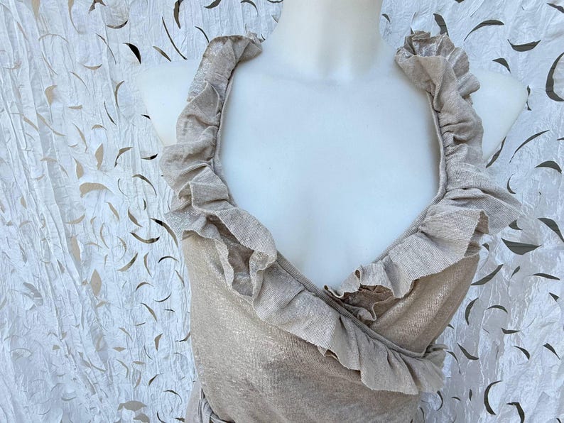 May include: A sleeveless beige top featuring a deep V-neckline and wide, ruffled trim that extends from the shoulders down to the waist. The fabric has a subtle metallic sheen, and the garment is displayed on a white mannequin torso.