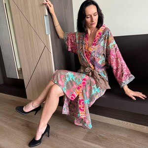 May include: A woman wearing a colorful patterned kimono-style robe with a brown sash. She is sitting on a brown bench with her legs crossed and wearing black heels.
