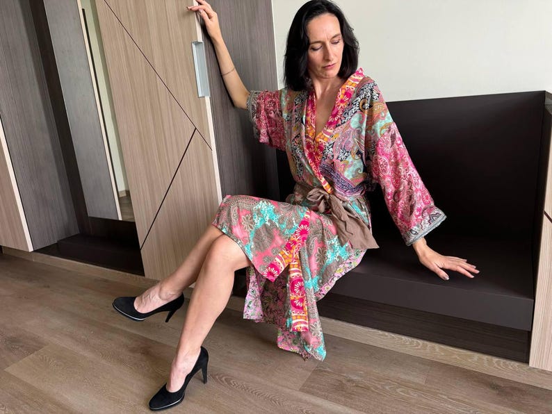 May include: A woman wearing a colorful patterned kimono-style robe with a brown sash. She is sitting on a brown bench with her legs crossed and wearing black heels.