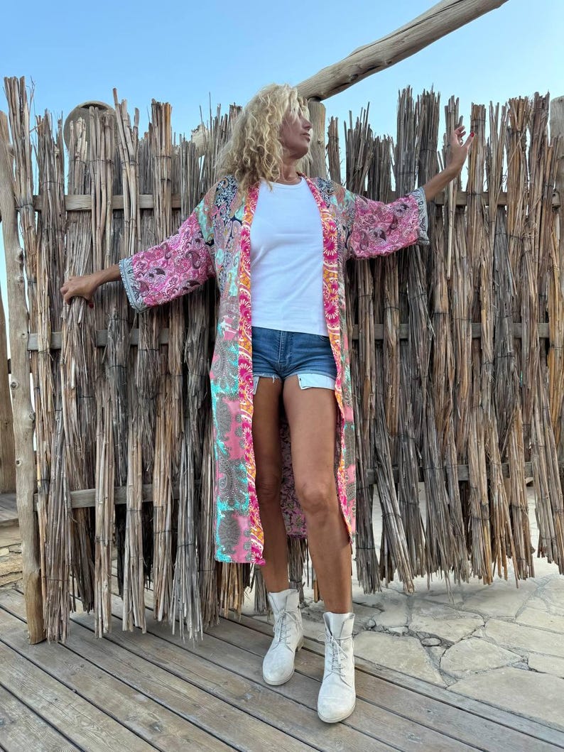 May include: A long, pink, and blue patterned kimono with a white top and denim shorts. The person is wearing white ankle boots and standing in front of a woven wood fence.