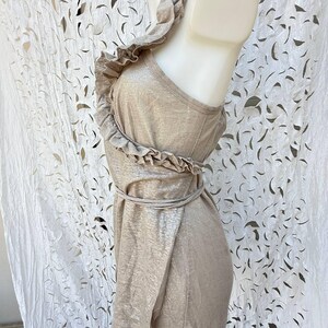 May include: A sleeveless metallic gold top featuring a ruffled trim that extends from the shoulder down the front. The garment includes a thin tie-waist detail and a textured, shimmering finish, displayed on a mannequin to show the side profile.