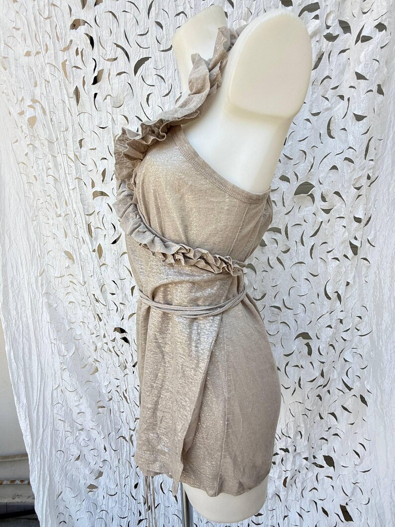 May include: A sleeveless metallic gold top featuring a ruffled trim that extends from the shoulder down the front. The garment includes a thin tie-waist detail and a textured, shimmering finish, displayed on a mannequin to show the side profile.