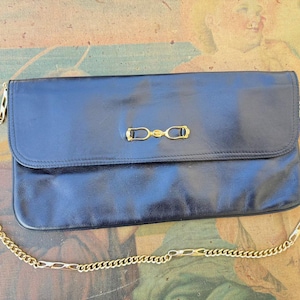 Könnte beinhalten: Eine rechteckige schwarze Clutch mit glatter Oberfläche und einem goldfarbenen Metallbeschlag im Trensen-Stil in der Mitte der Frontklappe. Die Tasche verfügt über einen abnehmbaren goldfarbenen Kettenschulterriemen mit länglichen Gliedern.