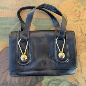 May include: A black leather handbag with gold-toned hardware and two circular accents. The bag has a top handle and a flap closure.