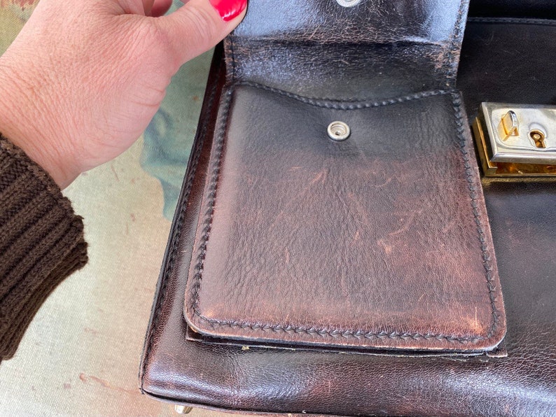 May include: A brown leather briefcase with a gold metal lock and a small pocket on the front. The briefcase is open and the inside is lined with a brown fabric.