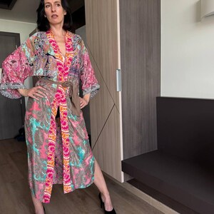May include: A woman wearing a long, colorful kimono with a floral pattern. The kimono has a sash tied at the waist and a long, flowing skirt. The woman is standing in a room with a wooden door and a bench.