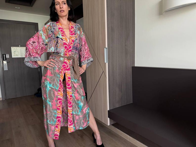 May include: A woman wearing a long, colorful kimono with a floral pattern. The kimono has a sash tied at the waist and a long, flowing skirt. The woman is standing in a room with a wooden door and a bench.