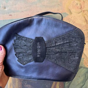 May include: A black leather handbag with a large bow detail. The bow is made of black suede and has a small gold label with the word "KENZO" on it.