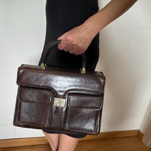 May include: A brown leather handbag with a top handle and two front pockets. The bag has a gold-toned clasp.