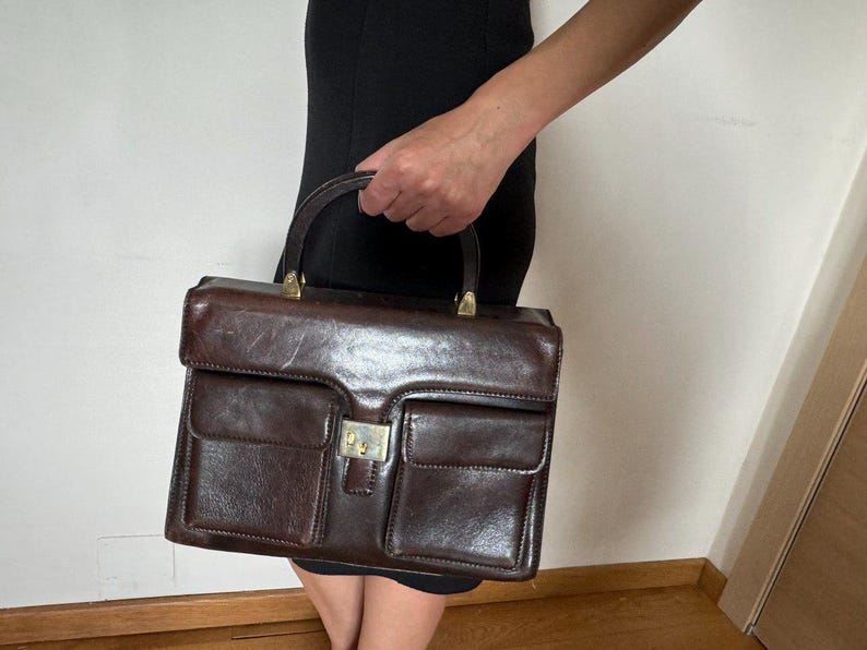 May include: A brown leather handbag with a top handle and two front pockets. The bag has a gold-toned clasp.