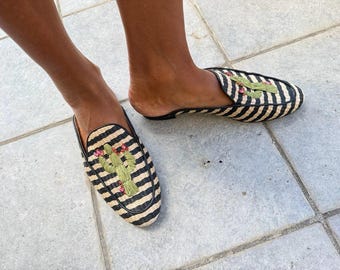 May include: A pair of slip-on mules featuring a woven straw texture with black and beige diagonal stripes. Each shoe is decorated with an embroidered green cactus motif accented with small pink details on the top of the rounded toe area.