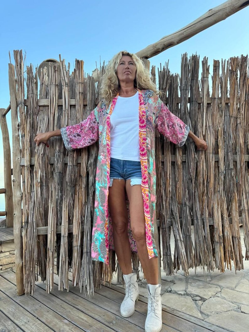 May include: A woman wearing a long, pink and blue floral kimono with a white t-shirt and denim shorts. She is standing in front of a woven wood fence and looking up.
