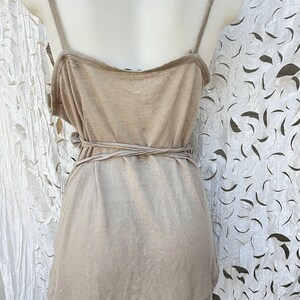 May include: A beige sleeveless tank top featuring thin shoulder straps and a wrap-around tie detail at the waist. The garment is displayed on a mannequin and has a simple, straight hemline, showcasing a solid, neutral-toned fabric construction.