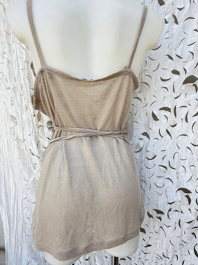 May include: A beige sleeveless tank top featuring thin shoulder straps and a wrap-around tie detail at the waist. The garment is displayed on a mannequin and has a simple, straight hemline, showcasing a solid, neutral-toned fabric construction.