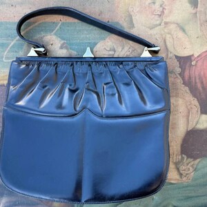 Könnte beinhalten: Eine schwarze Lederhandtasche mit einem silbernen Verschluss und einem plissierten Design. Die Tasche hat einen oberen Henkel und einen Klappenverschluss.