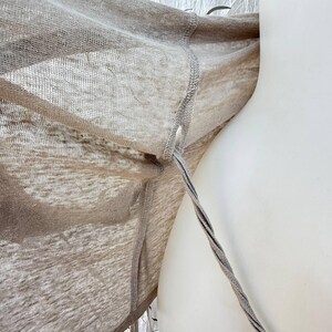 May include: A light beige, semi-sheer, lightweight knit garment featuring a visible seam along the side and a thin, twisted cord or strap extending from a small opening near the armhole area, showcasing a fine, open-weave texture throughout.