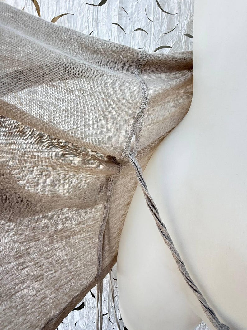 May include: A light beige, semi-sheer, lightweight knit garment featuring a visible seam along the side and a thin, twisted cord or strap extending from a small opening near the armhole area, showcasing a fine, open-weave texture throughout.