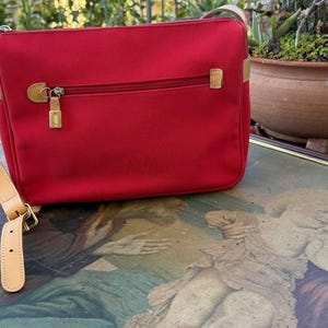 Può includere: Una borsa a tracolla in tela rossa con una tracolla e una fibbia in pelle marrone. La borsa ha una tasca con cerniera sul davanti.